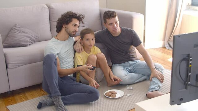 Happy gay parents and son eating cookies and watching movie at home, sitting on floor at TV and using remote control. Family and home entertainment concept.