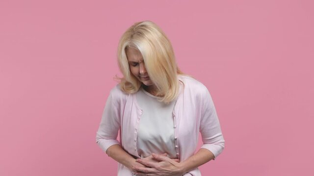 Sick ill elderly gray-haired blond woman lady 40s 50s years old in white blank casual t-shirt feel bad seedy put hands on abdomen with stomach-ache griping bellyache isolated on pastel pink background