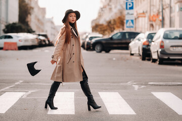 Fototapeta Stylish young woman in a beige coat in a black hat on a city street. Women's street fashion. Autumn clothing.Urban style obraz