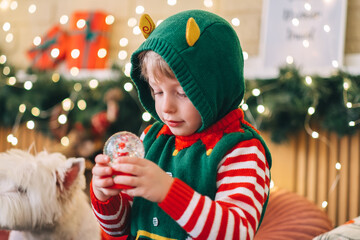 Happy new year concept. Happy blond child sits on the bed, opens a box and takes out a gift glass snow globe, the child smiles happily and is pleased with the gift.