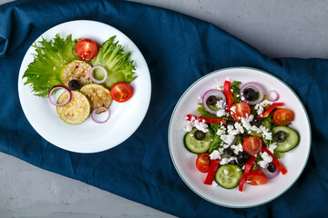 White plate with grilled zucchini with cherry tomatoes on lettuce and Greek salad on a blue napkin.