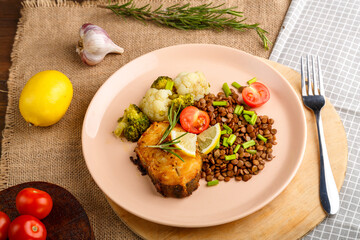 Baked fish with broccoli and lentils in a beige plate on a round stand next to tomatoes and lemon.