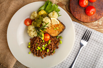 Baked fish with broccoli and lentils in a white plate with rosemary and lemon on a checkered napkin next to a fork.