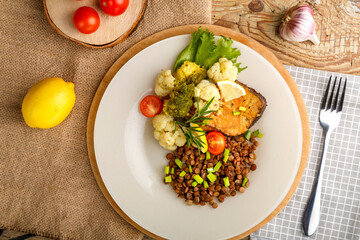 Baked fish with broccoli and lentils in a white plate with rosemary and lemon on a napkin and a round stand next to a fork and vegetables.