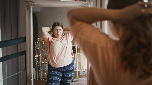 Plus size woman preening herself, making ponytail in front of mirror at home