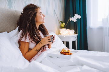 Woman have breakfast in bed in light hotel apartment or at home. Window light portrait young girl...
