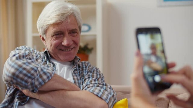 Handsome aged man posing on couch with wife taking picture on smartphone