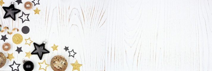 New Years Eve corner border of gold and black confetti stars, decorations and streamers. Top view over a white wood background.