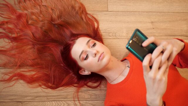 Redhead young pretty woman lie on a floor hair spreaded like a crown watch hold smartphone in social media