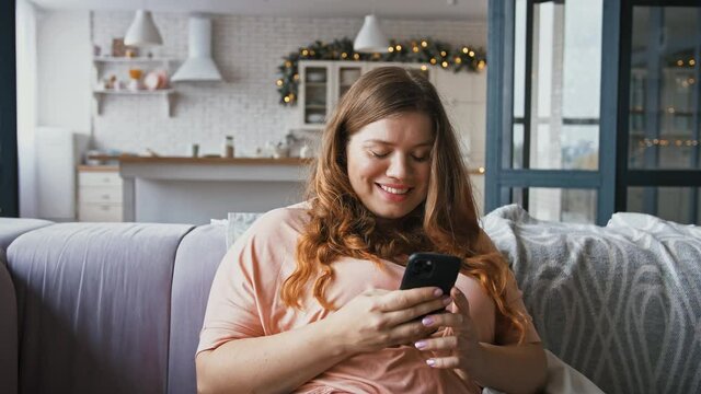 Relaxed young plus size woman using smartphone, surfing social media, sitting on sofa at home