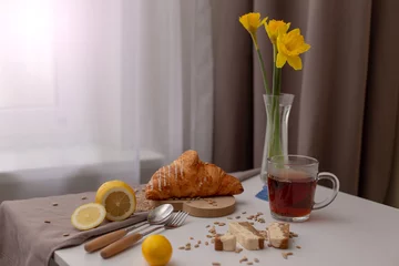 Tuinposter Breakfast with a cup of tea, croissants, lemons and yellow narcis in a glass vase © Natasha Zakharova
