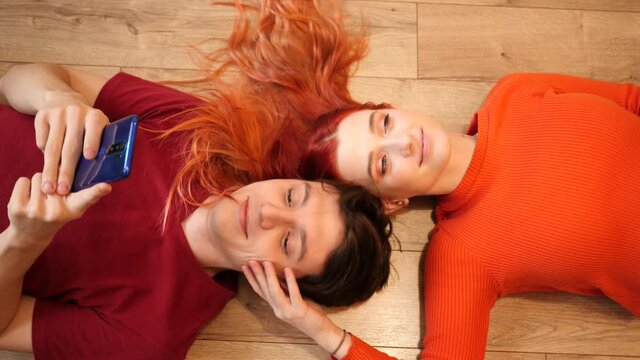 Young cute couple lie on floor man watch smartphone and girl redhead hair cherish his face tenderly