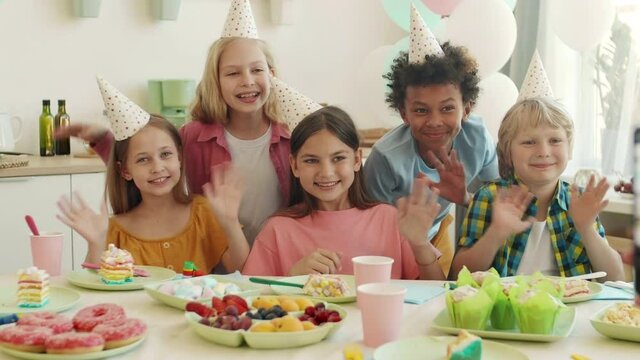 Medium of adult hand holding smartphone taking pictures of diverse school children wearing party hats sitting at table on birthday party. Boys and girls posing, smiling, waving hands on camera