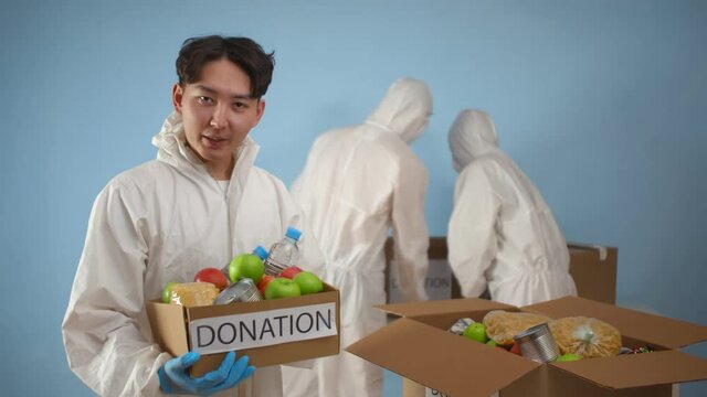 Portrait of asian man in protective suit holding box with donation food and talking on camera