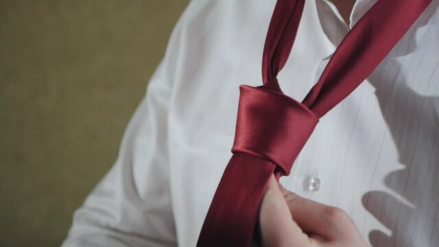 A man in a white shirt ties a red tie around his neck. learning tying a necktie