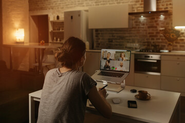 Back view of a female employee who is working remotely talking to her colleagues in a video conference on a laptop from home. A multiethnic business team on an online meeting in the evening.