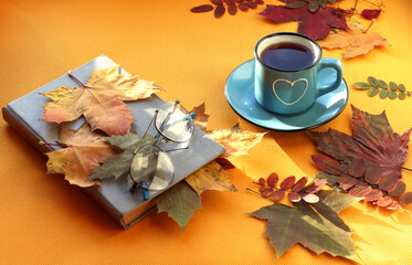 Naklejka na ściany i meble Blue mug with hot tea and a heart drawn, book, glasses, autumn foliage on a yellow background, place for text - the concept of leaving cozy autumn days
