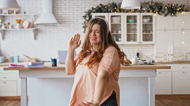 Cheerful overweight lady listening to music in wireless headphones and actively dancing at kitchen, fast motion