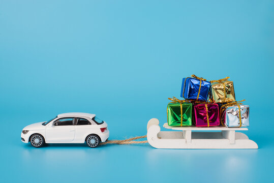 Christmas holidays concept. Close up photo of toy mini car carrying white wooden sledge full of small giftboxes isolated on blue background