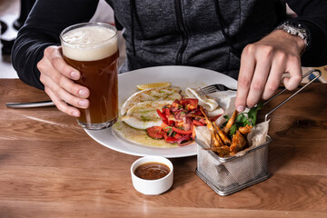 Grilled squid with tomato salad with red onion, roasted red mullet fish in deep-frying basket, glass of beer and sauce on wooden table. Men's hands holding a fork and a beer on a plate of squid.
