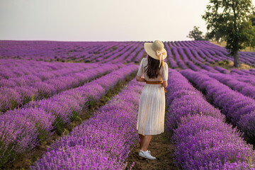 A young woman with long hair in a straw hat and dress stands in a lavender field. Photo from the...