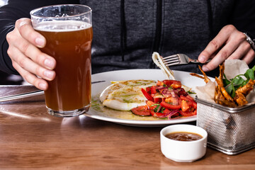 Grilled squid with tomato salad with red onion, roasted red mullet fish in deep-frying basket, glass of beer and sauce on wooden table. Men's hands holding a fork and a beer on a plate of squid.
