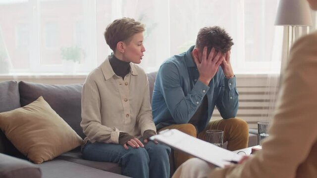 Medium over shoulder of Caucasian married couple sitting on couch in front of unrecognizable red-haired female family psychologist. Woman talking, criticizing, looking at annoyed husband rubbing face