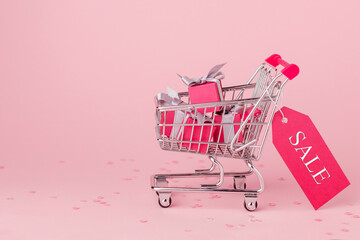 Small metal trolley, mini cart filled with pink gift paper boxes on pink background. Preparing for celebration, buying gifts, sale. Market concept.