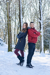 Fototapeta na wymiar Young couple in love, enjoying winter weather outside in park forest. Brunette man in red jacket and woman in black, standing back to back,smiling. Christmas and new year holidays joy. Romantic date.