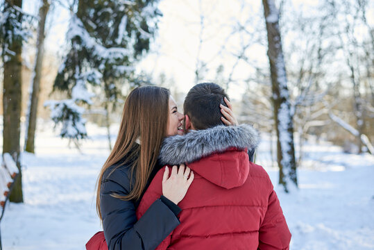 Young couple in love, enjoying winter weather outside in park forest. Brunette man in red jacket and woman in black, hugging, looking at each other. Christmas and new year holidays joy. Romantic date.