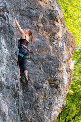 Fototapeta premium happy beautiful caucasian woman with long brown hair rock climbing, flexing her muscles on a sunny day in the mountains, strongly holding on to cliffs. Healthy outdoor sports and activities in nature