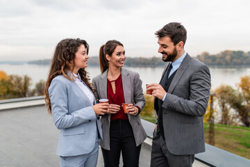 Three business people standing on the roof of modern building. They are drinking coffee, talking and smiling. Beautiful river landscape in the background.