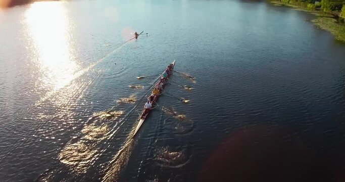 Drone shot on a sports canoe driven by a team of men and women sailing along a calm river with the sun reflected in the water.