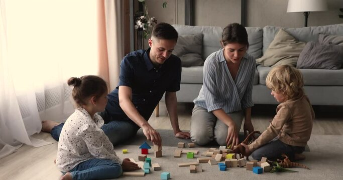 Happy young parents play with daughter and son preschool children with wooden blocks and dino toys develop kids together at weekend at modern warm home. Homeowners leisure pursuit, free time concept