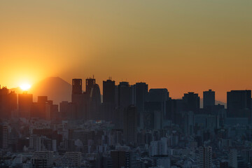 Naklejka premium 夕陽が沈むオレンジの空を背景にシルエットになって見える富士山と新宿のビル群