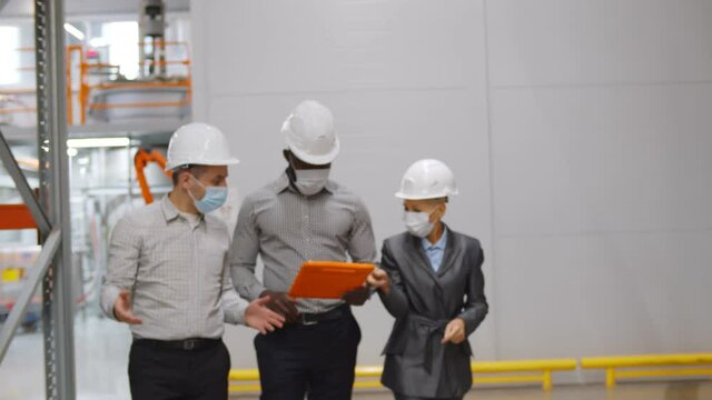 Engineer manager in safety mask and helmet giving tour two businessmen to inspect factory warehouse