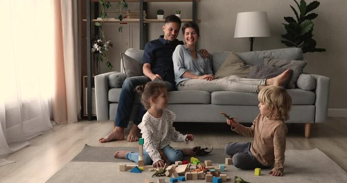 In modern living room gathered family with small kids, couple rest on sofa their son and daughter play game together with wood blocks and dino toys set. Family leisure at comfort house owners concept