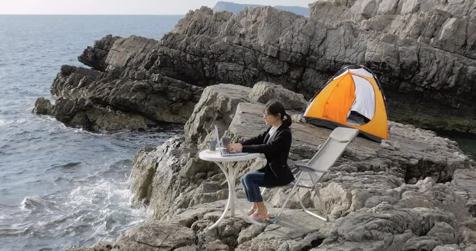 Female Business woman sitting on the rock above the sea and typing on laptop. Freelancer working from the beach. Working from distance concept. 