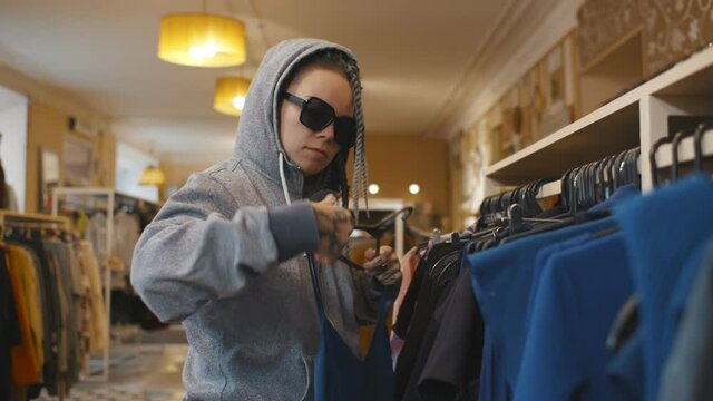 Side view of young woman in sunglasses and hood stealing clothes from store