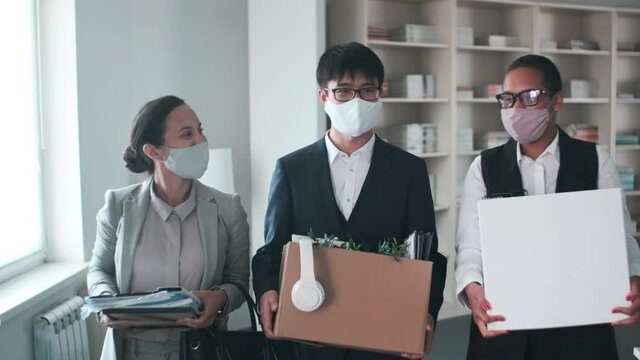 Group of three cheerful multi-ethnic people wearing protective masks bringing their belongings in box to office after working at home period and talking to each other