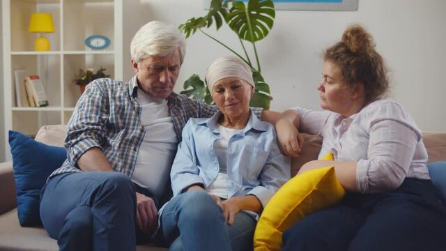 Senior cancer woman with headscarf sitting on couch with husband and daughter