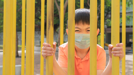 The lovely Asian boy with the surgical mask are holding the yellow playground toy rails or iron gate. The funny face kid is playing at the outdoor play area. 