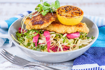 A serving of Chicken Schnitzel in a bowl with fresh coleslaw, and a fried lemon wedge on top.
