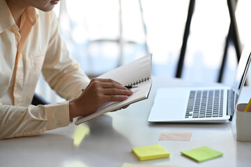 Cropped shot of a male hand taking notes on notebook while sitting in front of laptop at office.