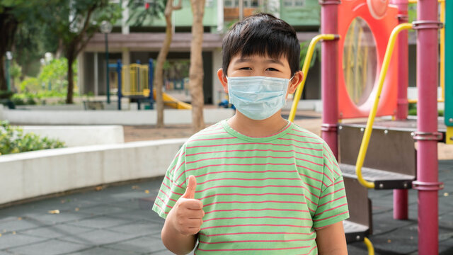 One Asian boy is wearing the disposal mask and showing a thumb up gesture at the park playground with no-one staying or surrounding him. Kid and the virus prevention/safety protection concept.