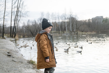 Fototapeta na wymiar a little boy stands by the lake in winter clothes, looks at the floating ducks, a walk with a child