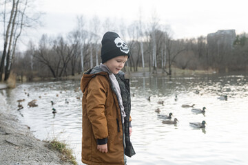Fototapeta na wymiar a boy slyly looks at the camera, on the street in winter clothes, a black hat and a mustard jacket, a little handsome man, against the background of floating ducks, winter nature, autumn fairy tale