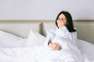 Brunette woman in white robe yawning with covered mouth in comfy bed with side light from left