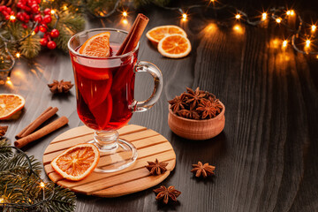 Obraz na płótnie Canvas Mulled wine in a beautiful glass with orange and cinnamon on a dark background with fir branches, garland and red berries. Nearby are orange slices, cinnamon, anise and spruce branches. Copy space.