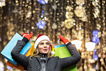 A woman with long hair and a Santa hat near the window of a city store with purchases in colorful, paper bags. New year's shopping